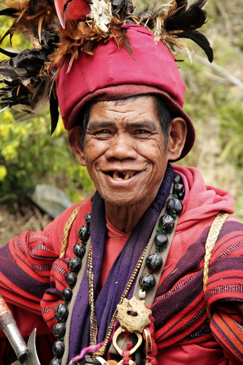 Banaue, Philippines- January 25, 2013: Head shot portrait of a native ifugao man in a traditional warrior outfit on the road to the Banaue rice terraces in the province of Ifugao, Philippines. The man is dressed in red, has feathers on his head and traditional jewellery around his knock.