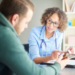 Vrouwelijke huisarts in gesprek met een cliënt die naar een buisje pillen kijkt.